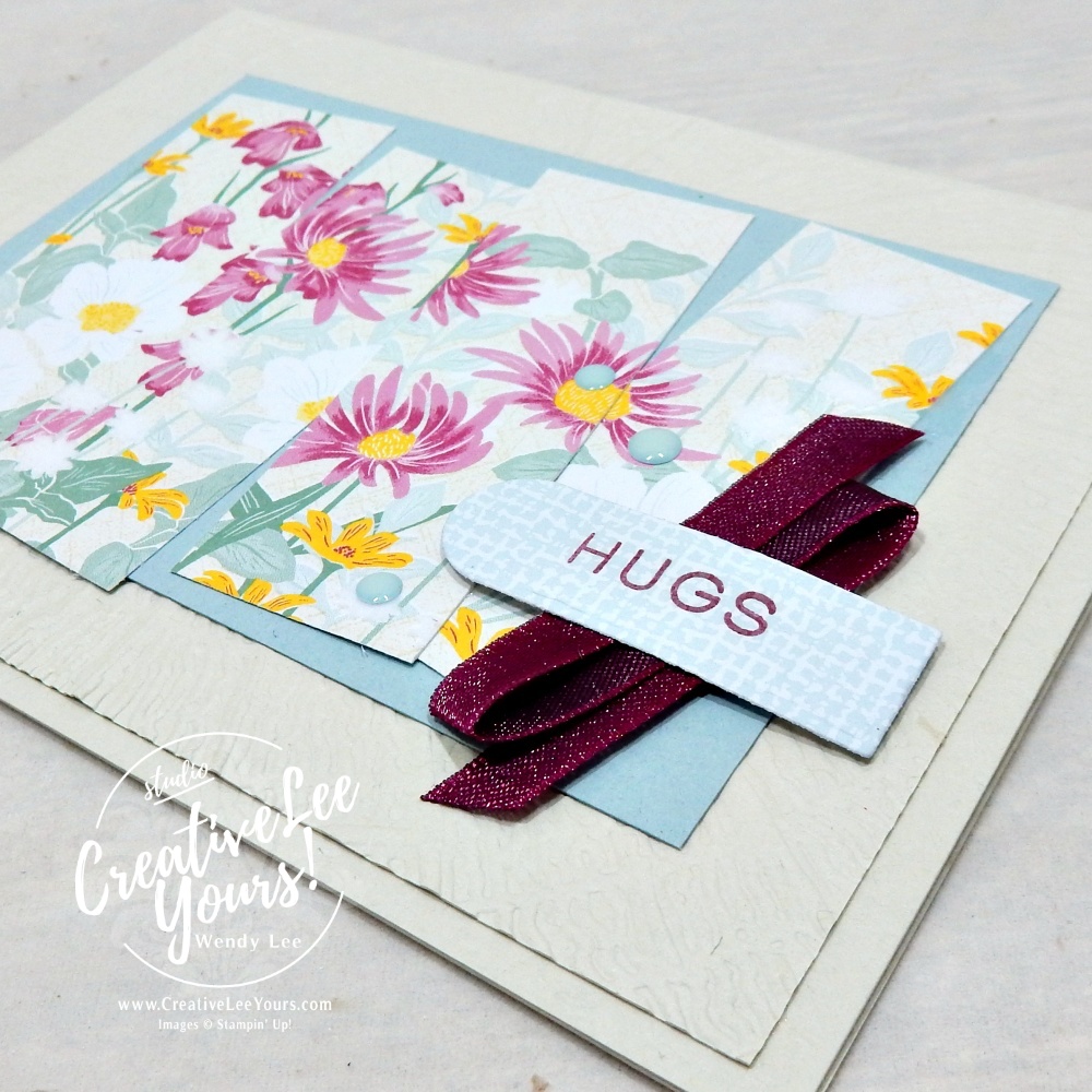 Close-up of embossed tree ring background with layered designer paper strips, satin ribbon, scalloped die-cut sentiment, and Moody Dots embellishments.